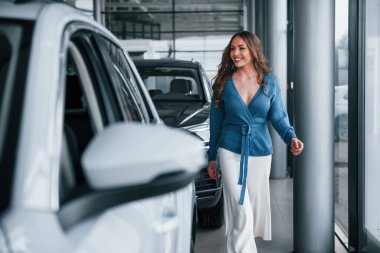 Positive woman in blue shirt walks near brand new car. In auto salon.