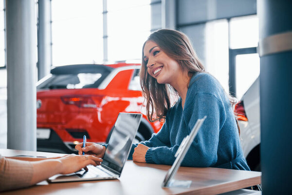 Cheerful woman sits with assistant in car shop and buying new vehicle.