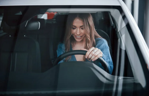 Front view of positive woman in blue shirt that sits inside of new brand new car. In auto salon or airport.