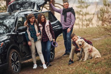 Happy family have fun with their dog near modern car outdoors in forest.