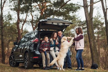 Happy family have fun with their dog near modern car outdoors in forest.