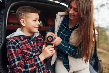 Kız kardeşi, erkek kardeşi için ormanda sıcak su dökerken arabanın arkasında oturuyor..