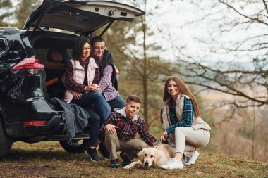 Mutlu bir aile ormanda, modern arabalarının yanında oturmuş köpekleriyle eğleniyor..