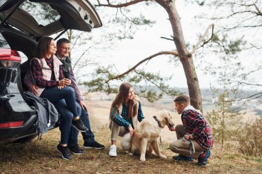 Happy family sitting and having fun with their dog near modern car outdoors in forest.