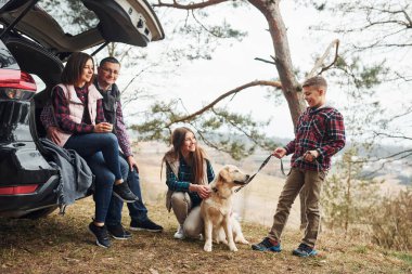 Happy family sitting and having fun with their dog near modern car outdoors in forest.