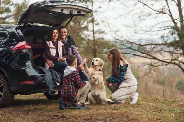 Happy family have fun with their dog near modern car outdoors in forest. clipart
