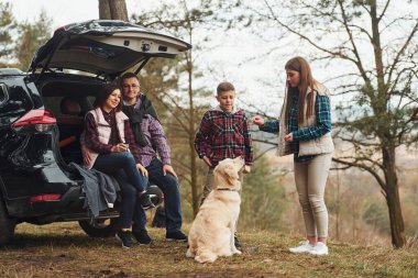 Happy family have fun with their dog near modern car outdoors in forest.