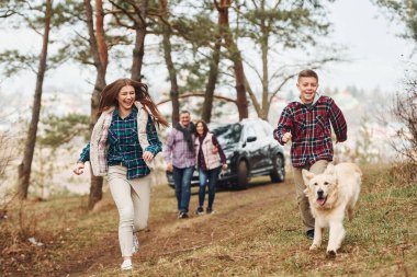 Happy family runs and having fun with their dog near modern car outdoors in forest. clipart