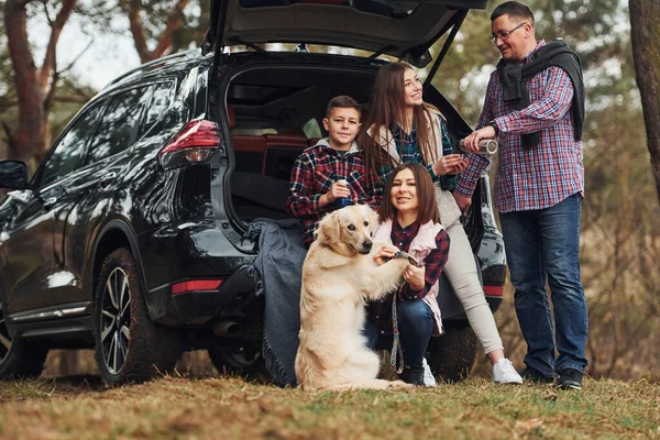 Happy family have fun with their dog near modern car outdoors in forest.