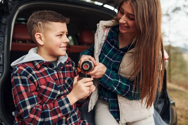 Kız kardeşi, erkek kardeşi için ormanda sıcak su dökerken arabanın arkasında oturuyor..