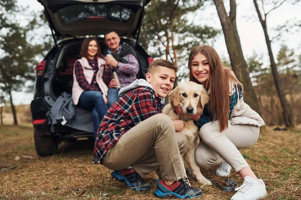 Happy family sitting and having fun with their dog near modern car outdoors in forest.