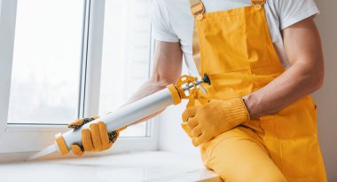 Handyman in yellow uniform works with glue for window indoors. House renovation conception.