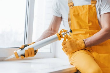 Handyman in yellow uniform works with glue for window indoors. House renovation conception.