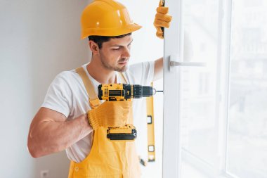 Sarı üniformalı tamirci otomatik tornavida kullanarak yeni pencere kuruyor. Ev yenileme konsepti.