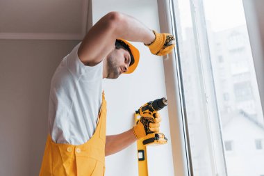 Handyman in yellow uniform installs new window by using automatic screwdriver. House renovation conception.
