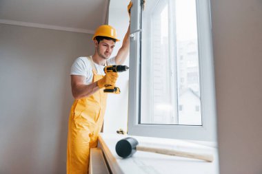 Handyman in yellow uniform installs new window by using automatic screwdriver. House renovation conception.