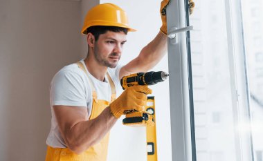 Handyman in yellow uniform installs new window by using automatic screwdriver. House renovation conception.