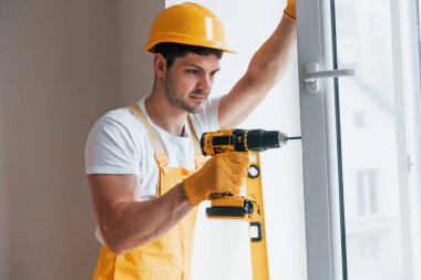 Sarı üniformalı tamirci otomatik tornavida kullanarak yeni pencere kuruyor. Ev yenileme konsepti.
