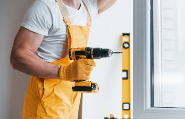 Sarı üniformalı tamirci otomatik tornavida kullanarak yeni pencere kuruyor. Ev yenileme konsepti.