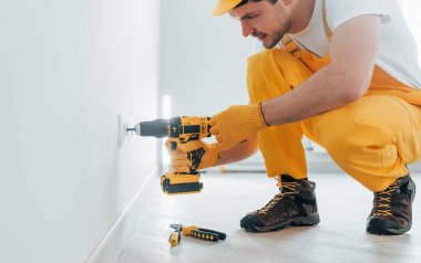 Handyman in yellow uniform works with electricity and installing new socket by using automatic screwdriver. House renovation conception.