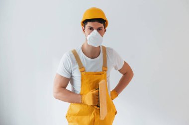 Handyman in yellow uniform and protective mask standing against white wall. House renovation conception.