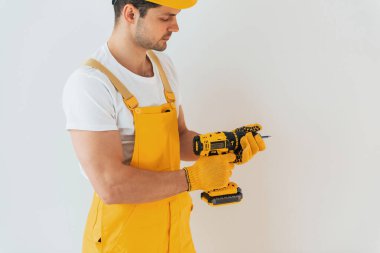 Handyman in yellow uniform standing against white wall with automatic screwdriver. House renovation conception.
