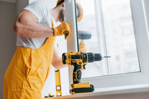 Handyman in yellow uniform installs new window by using automatic screwdriver. House renovation conception.