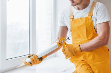 Sarı üniformalı tamirci içeride pencere için yapıştırıcı kullanıyor. Ev yenileme konsepti.