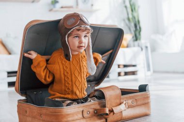 Little boys in retro pilot costume have fun and sitting in suitcase indoors at daytime.