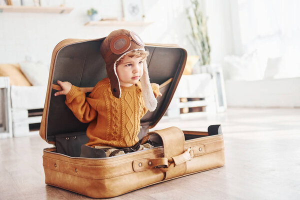 Little boys in retro pilot costume have fun and sitting in suitcase indoors at daytime.