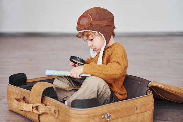Little boys in retro pilot costume have fun and sitting in suitcase indoors at daytime.