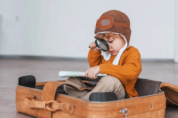 Little boys in retro pilot costume have fun and sitting in suitcase indoors at daytime.