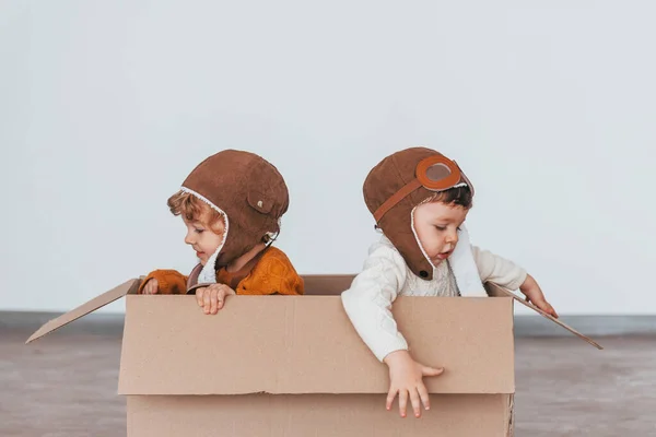 Two little boys in retro pilot costumes have fun and sitting in paper box indoors at daytime.