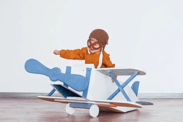 Little boy in retro pilot uniform having fun with toy plane indoors.