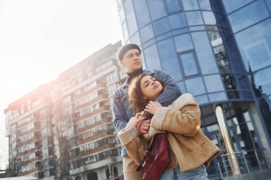Cheerful couple in casual warm clothes have a walk outdoors in the city near business building.