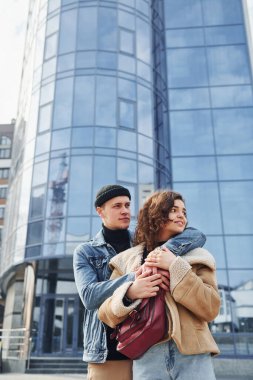 Cheerful couple in casual warm clothes have a walk outdoors in the city near business building.