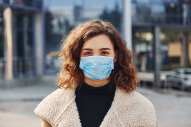 Karantina zamanında iş yerinin yakınında duran koruyucu maskeli kıvırcık saçlı genç kızın portresi. Koronavirüs kavramı.