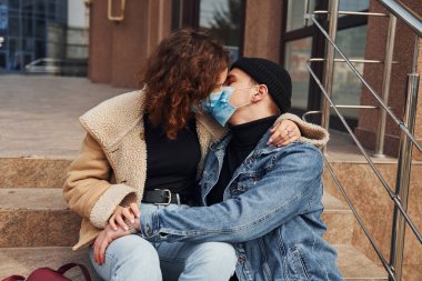 Karantina zamanında iş binasının yakınında koruyucu maskeli bir çift birbirini öpüyor. Koronavirüs kavramı.