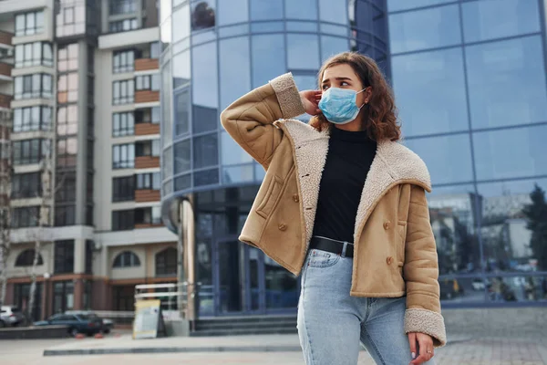 Karantina zamanında iş yerinin yakınında duran koruyucu maskeli kıvırcık saçlı genç kızın portresi. Koronavirüs kavramı.