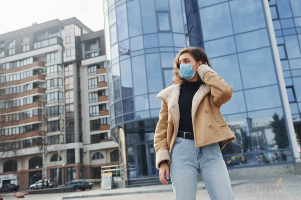 Karantina zamanında iş yerinin yakınında duran koruyucu maskeli kıvırcık saçlı genç kızın portresi. Koronavirüs kavramı.