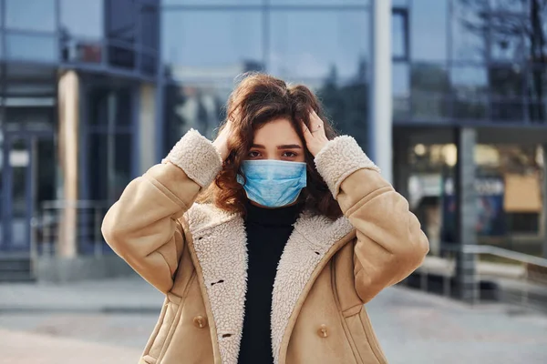 Portrait of young girl with curly hair in protective mask that standing outdoors near business building at quarantine time. Conception of coronavirus.