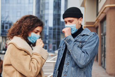 Koruyucu maskeli bir çift karantina zamanında iş binasının yakınında dışarıda öksürüyorlar. Koronavirüs kavramı.