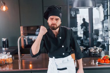 Profesyonel aşçı üniforması içinde masanın yanında duruyor ve mutfakta kameraya poz veriyor..