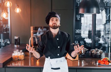 Profesyonel aşçı üniforması içinde masanın yanında duruyor ve mutfakta kameraya poz veriyor..