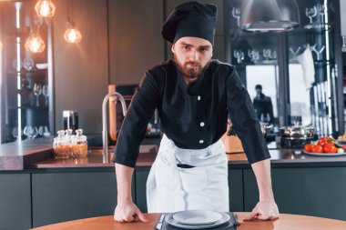 Profesyonel aşçı üniforması içinde masanın yanında duruyor ve mutfakta kameraya poz veriyor..