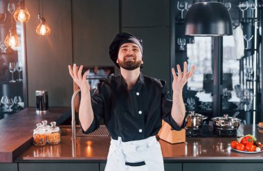 Profesyonel aşçı üniforması içinde masanın yanında duruyor ve mutfakta kameraya poz veriyor..