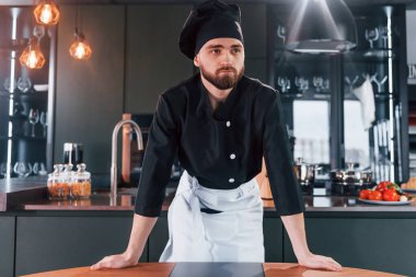 Profesyonel aşçı üniforması içinde masanın yanında duruyor ve mutfakta kameraya poz veriyor..