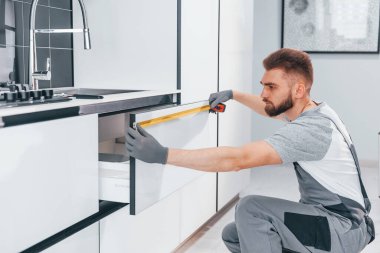 Young professional plumber in grey uniform measuring table by using meter on the kitchen.