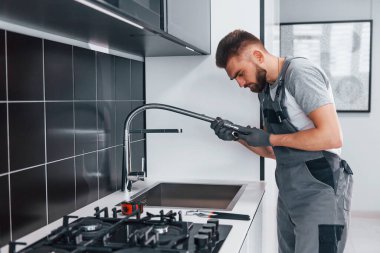 Young professional plumber in grey uniform fixing water tap on the kitchen.