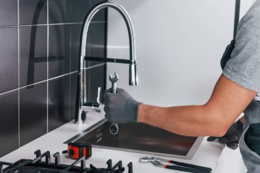 Close up view of young professional plumber in grey uniform holding wrench in hand on the kitchen.
